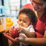 Mère souriante tenant son bébé sur les genoux pendant le repas, illustrant le soutien familial et la pension alimentaire.