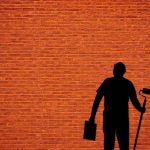 Un homme en noir tient un rouleau et un seau contre un mur rouge